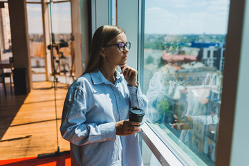 A young female freelancer in glasses is standing with coffee near a large window and looking at the city. A girl in a shirt and shorts stands in a cafe by the window
