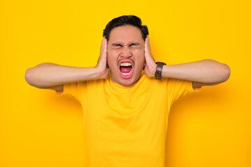 Fototapeta na wymiar Angry young Asian man in casual t-shirt screaming with closed eyes and open mouth, covering ears with hands isolated on yellow background. People lifestyle concept