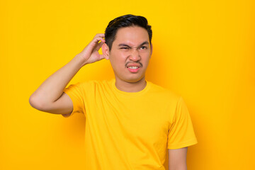 Confused young Asian man in casual t-shirt touching his head with hand, having problem isolated on yellow background. People lifestyle concept