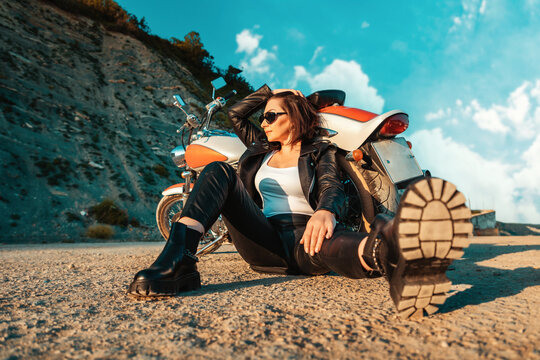 A cool independent woman in a leather motorcycle outfit poses sitting on the ground near a motorcycle. Low angle view. The concept of the motorcycle travel and feminism