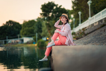 A young smiling woman in summer clothes and straw hat sitting at river embankment. Summer vacation and relax