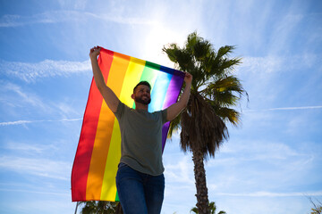 Handsome young gay man holding the gay pride flag in the wind, blue sky in the background. The flag...