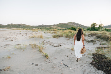 Unrecognizable woman wearing a white dress and holding a hat while walking outdoors in nature.