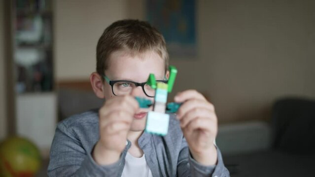 Caucasian child in glasses twists colored toy built from constructor bricks in front of his face. Boy carefully examines his toy product, a toy man. Focus on the child face.