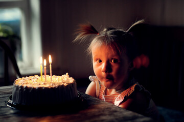 Funny toddler girl with two ponytails will conceive 3 candles on birthday cake, dark style and soft...