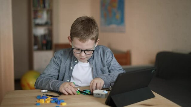 Caucasian boy with glasses creates his own model of programmable robot from plastic constructor. Child sits at table, clicks on screen in tablet and connects the details of the toy.