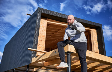 Naklejka na ściany i meble Male developer building wooden frame house on pile foundation. Portrait of bald man sitting on terrace on construction site, inspecting quality of work.