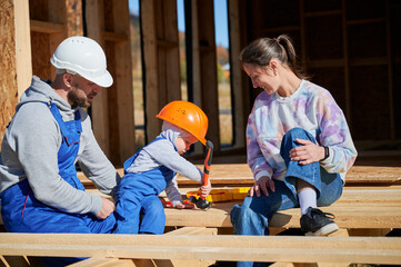 Father, mother and son building wooden frame house. Toddler boy helping his daddy, while woman...