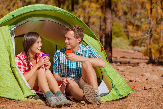 Camping couple drinking water in tent smiling happy outdoors in forest enjoying sun at looking at view. Happy multiracial couple relaxing after outdoor activity hiking. Asian woman, Caucasian man
