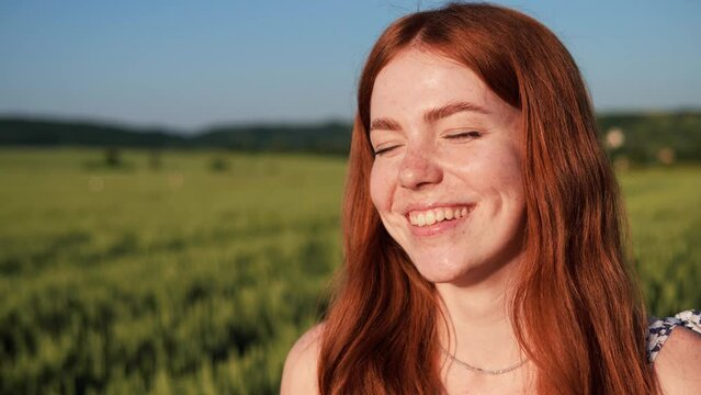 Portrait of attractive ginger woman with freckles enjoys nature at sunset. Dreamer red hair young beautiful girl looking at camera breathes fresh air. Female standing in wheat field with blue sky.