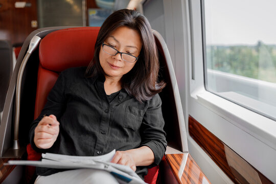 Train commute mature Asian businesswoman working on business trip commuting to work. Happy woman sitting by window in luxury first class car reading papers
