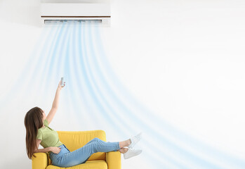 Young woman switching on air conditioner while sitting in armchair near white wall