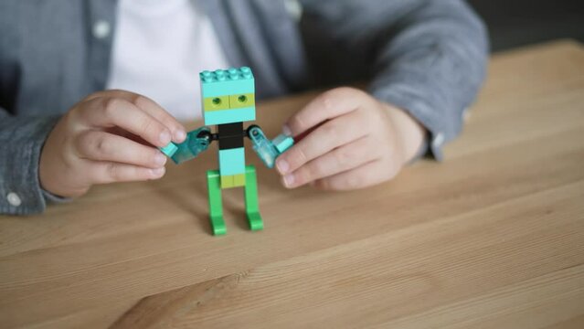 Child attaches the last bricks to the toy from constructor. The boy is playing with a ready-made toy man, a colorful hero is dancing on a wooden table, close-up.