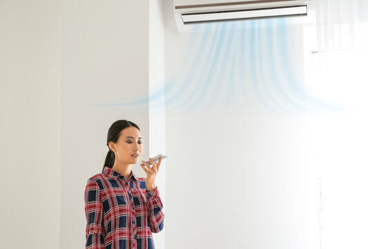 Young Asian Woman With Mobile Phone Adjusting Air Conditioner. Modern Technology And Smart Home Automation