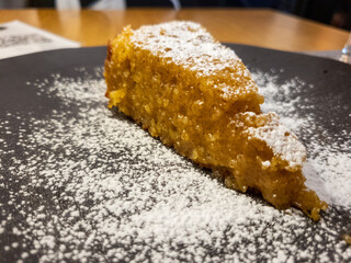 A slice of almond cake covered on powdered sugar on a black plate