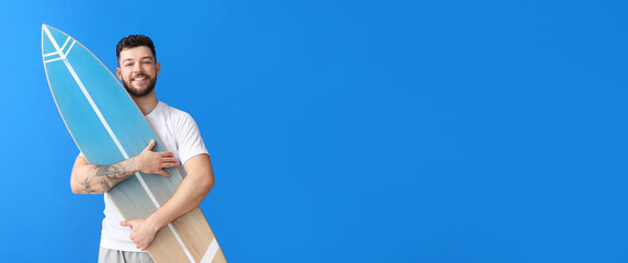 Happy young tattooed man with surfboard on blue background with space for text