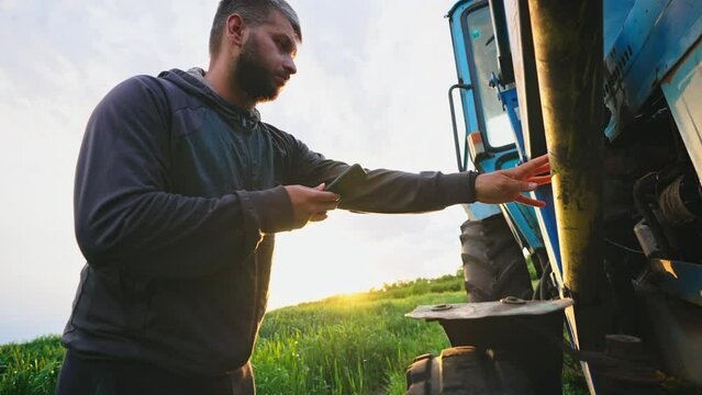 Man in field with phone in his hands stands by his tractor and knocks on wheel, checking condition of machine to diagnose serviceability of equipment. Running a small business selling grown products.