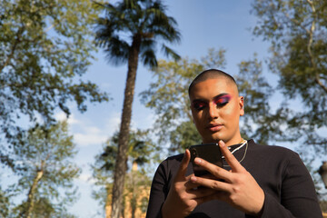 Portrait of non-binary person, young and South American, heavily make up, consulting social networks on his cell phone. Concept queen, lgbtq+, pride, queer.