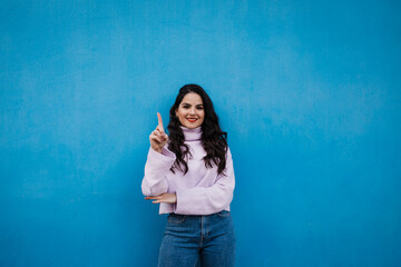 Smiling young beautiful woman showing number one with index finger in front of blue wall