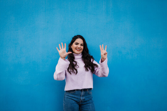 Smiling young beautiful woman showing number 8 in front of blue wall