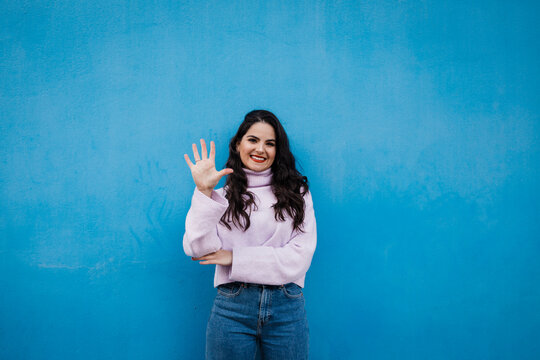 Smiling young beautiful woman showing number 5 in front of blue wall