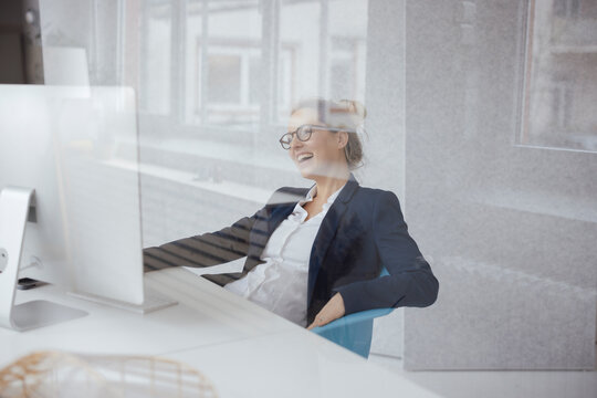 Happy businesswoman with desktop PC at desk seen through glass