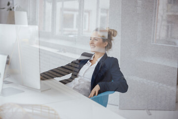 Happy businesswoman using desktop PC at desk seen through glass