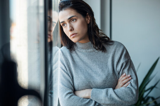 Depressed woman standing with arms crossed looking through window at home