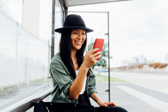 Smiling young woman surfing net through smart phone sitting at bus stop