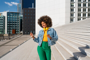 Happy Afro woman wearing wireless headphones enjoying dance by steps on sunny day