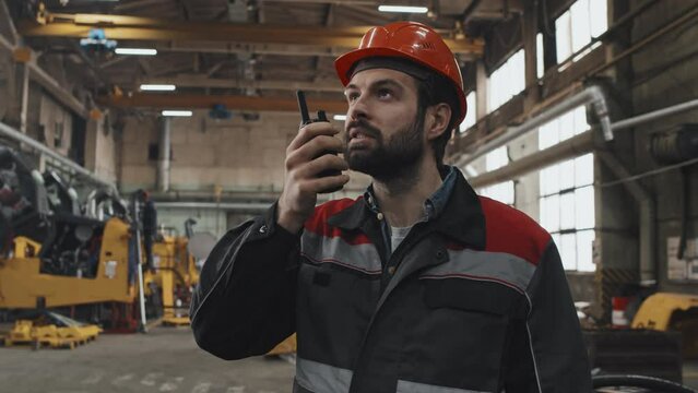 Medium close-up of Caucasian bearded male plant manager talking into walkie talkie at factory in afternoon