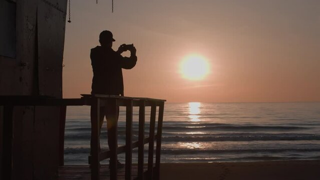 Male traveler in cap and clothes makes panorama shoot of amazing sunset. Silhouette of adult man standing on observation deck, calm sea, clear sky, small waves, empty beach on background. Slow motion