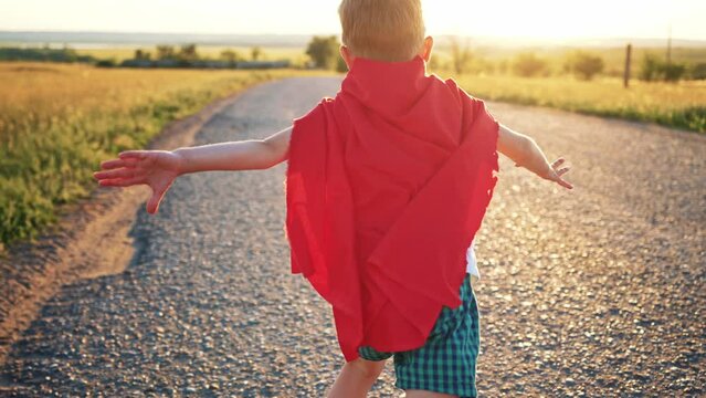 Boy Plays Superhero. Happy little boy are playing superhero. Kid run across road in red cloak at sunset time. Pretty child superhero hero in red cloak in nature. Freedom power concept.