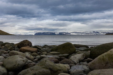 Pebbles and rocks on the shore of Icelandic coastline with snow covered mountain range behind fiord’s body of water