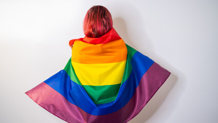 Young red-haired woman stands with her back against a white background and holds the lgbt flag