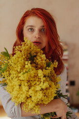 Red hair woman posing for the camera while holding a bouquet  of yellow flowers. There are books in the background, stacked on the floor. 
