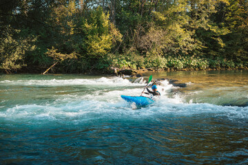 Young teenager cruising down whitewater rapids in a blue kayak, beautiful river nature on a sunny summer day, handheld shot.