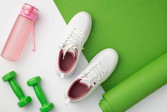 Fitness concept. Top view photo of white sneakers pink bottle of water green exercise mat and dumbbells on isolated white background