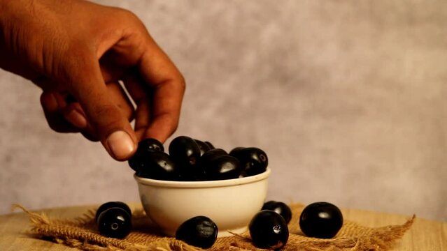 Man eating jamun fruit on white bowl with dark background, blueberries picking on hand, berry pick up man