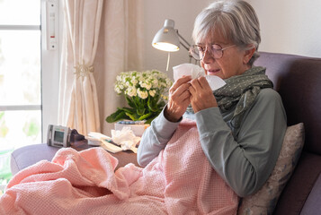Older adult caucasian woman with severe cold and fever symptoms such as seasonal flu or pollen allergy - woman on sofa at home with blanket