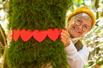 Smiling elderly woman hugging a moss covered tree trunk in the woods with a wreath of small red hearts - earth day concept. People save the planet from deforestation