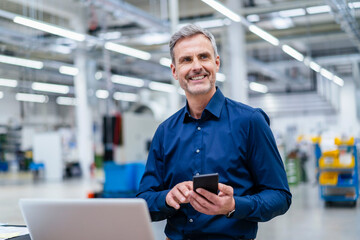 Mature businessman using laptop and smartphone in factory