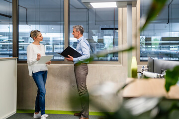Businessman with clipboard and businesswoman with digital tablet talking in factory office