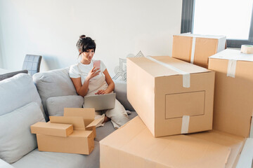 Woman using mobile phone sitting with laptop and cardboard boxes in living room