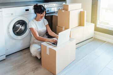Happy woman using laptop on cardboard box by washing machine in utility room