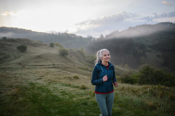 Fototapeta na wymiar Senior woman jogging in nature on early morning with fog and mountains in background.