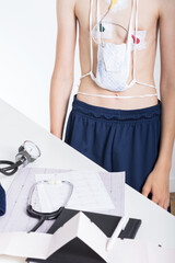 Standing teenager on white background with three electrodes on the chest near the table with electrocardiogram and stethoscope. Heart electrocardiogram or monitoring using Holter for young patient