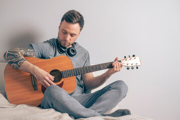 Man playing acoustic guitar at home. Serious, focused face expression. Singing serenade for girlfriend. Handsome young man is sitting on the bed and composing music, using laptop and headphones
