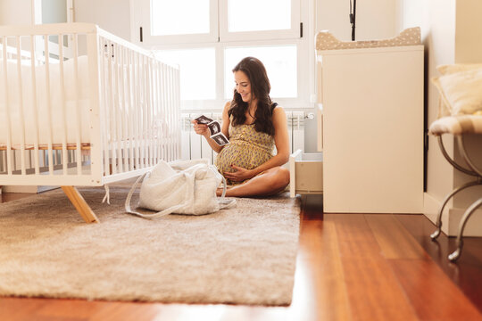 Smiling pregnant woman with ultrasound scans sitting by crib at home