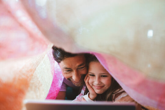 Happy daughter and father using tablet PC lying under blanket